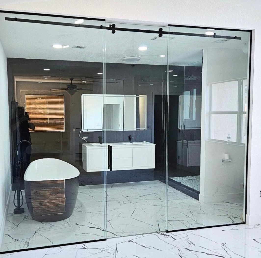 Floor-to-ceiling glass wall on a black sliding track enclosing a marble bathroom with a freestanding tub